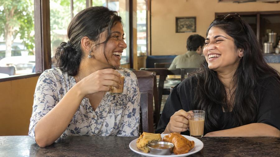 Illustration: two friends laughing over glasses of chaya and snacks at a café